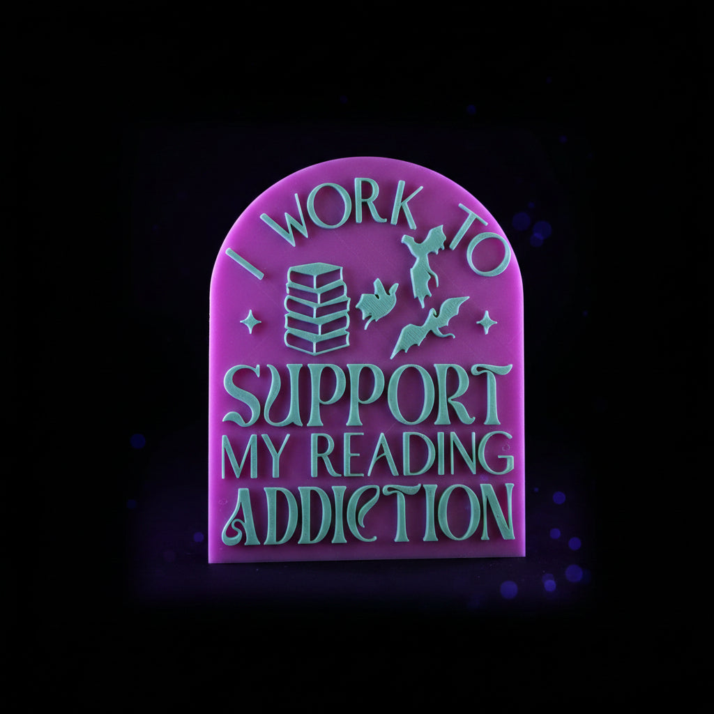 3D printed I Work To Support My Reading Addiction arched bookshelf sign in pink with green raised lettering, bats, and book stack - flat lay on printer bed showing sign and base