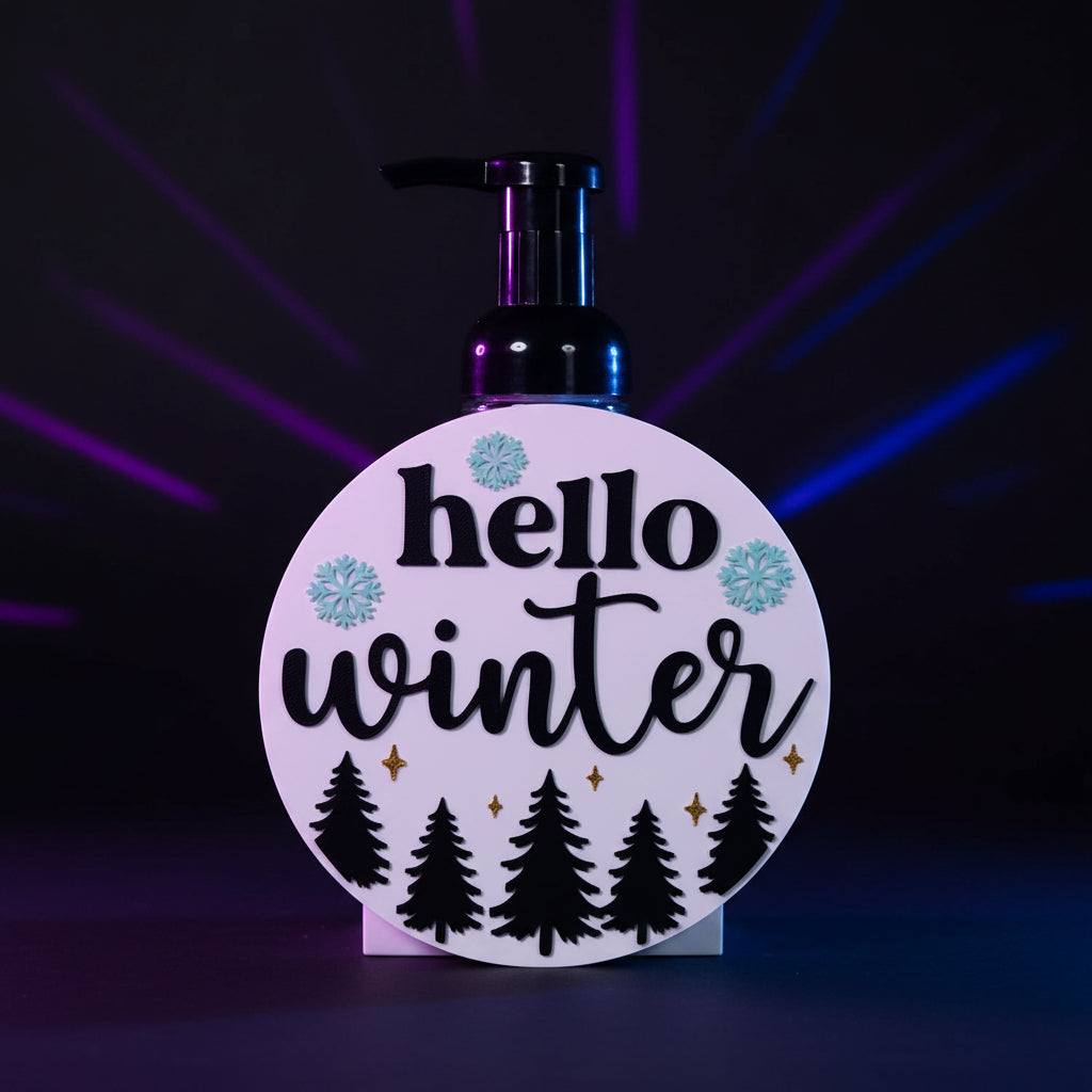 3D printed Hello Winter BBW interchangeable soap cover with raised black text, icy blue snowflakes, gold stars, and pine trees on white round base - front view on foaming dispenser